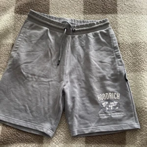 Grå shorts från Hoodrich med tryck - Snygga grå shorts från Hoodrich med tryck och broderade detaljer på benen. Modellen har elastisk midja med snörning och flera fickor. Perfekta för chill dagar eller streetwear-stil. Mjukt bomullsmaterial för skön känsla.