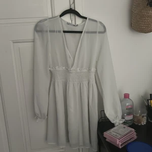 Vit omlottklänning från 157, XS - Supersöt vit kortklänning från 157 med omlott-v-ringning, långa ärmar och smockad midja med volangdetalj. Kolla sista bild för se fläck, aldrig använd bara testad.