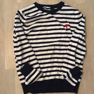 Randig tröja från Ami Alexandre Mattiussi - Säljer en snygg långärmad tröja från Ami Alexandre Mattiussi med marinblå och vita ränder. Tröjan har en broderad röd hjärtlogga på bröstet och ribbade muddar vid ärmslut och hals. Perfekt för dig som gillar klassisk stil med en twist.