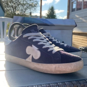 Mörkblå sneakers från The Gallerist OG Model - Snygga mörkblå sneakers från The Gallerist med vit klöverdetalj på sidan. Skorna har vit snörning, vit hälkappa och svart sula. Ovandelen är i mocka för en cool och lite lyxig känsla. Perfekta för dig som vill sticka ut med en unik detalj. Skorna är OG modellen så det går inte att få tag på dem. Skorna är använda ett par gånger men är ändå i  mycket bra skick. Boxen är inte i bästa skick.