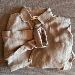 Olivgrön  linneskjorta från Kappahl - Snygg Olivgrön skjorta i linne från Kappahl med klassisk krage, bröstficka och knäppning framtill. Skjortan har långa ärmar som enkelt kan vikas upp för en mer avslappnad look. Perfekt för dig som gillar stilrena och enkla plagg. Hör av er vid intresse/ frågor!