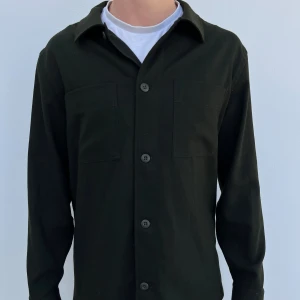 Mörkgrön overshirt från Massimo Dutti - Stilren svart overshirt från Massimo Dutti med klassisk krage, två bröstfickor och knäppning framtill. Skjortan är långärmad och tillverkad i ett kraftigare bomullstyg som ger en clean och modern look. Perfekt att bära öppen över en t-shirt.