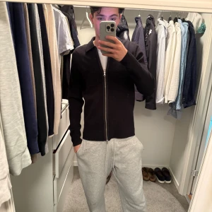 Samsøe tröja - Svart, stilren kofta med hel dragkedja framtill och klassisk krage. Modellen är långärmad och har en slim passform som sitter snyggt på kroppen. Perfekt för dig som gillar en clean look och vill ha något enkelt att slänga på sig. Materialet känns mjukt och bekvämt.