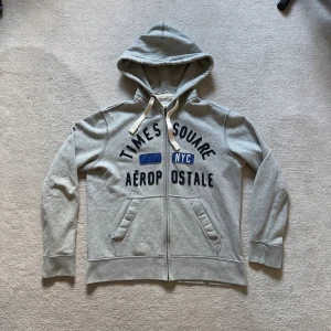 Vintage zip hoodie från Aeropostale NYC - Grå vintage zip hoodie från Aeropostale med tryck 'Times Square NYC' i blått och svart framtill. Tröjan har huva med snörning, dragkedja och två fickor. Perfekt för chill dagar och streetstyle. Materialet är mjuk bomullsmix som känns skönt mot huden. Storlek M men passar S.