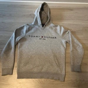Grå hoodie Tommy Hilfiger M - Snygg grå hoodie från Tommy Hilfiger i ekologisk bomull. Klassisk logga och text på bröstet, huva med dragsko och magficka. Perfekt för chill dagar och enkel att matcha med jeans eller joggers.