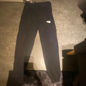 Svarta mjukisbyxor från The North Face - Svarta mjukisbyxor från The North Face med logga på vänster lår. Byxorna har en slim passform och är tillverkade i mjukt bomullsmaterial. Perfekta för chill eller träning, med elastisk midja och muddar vid bensluten.