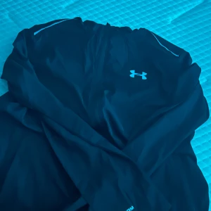 Svart vindjacka från Under Armour - Svart vindjacka från Under Armour med reflekterande logga på bröstet och stor text på ryggen. Jackan har dragkedja framtill och är tillverkad i lätt polyester. Perfekt för träning och aktiva dagar, med långärmad design och sportig look.priset går och diskuteras