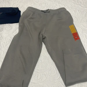 Grå mjukisbyxor från Hoodrich - Snygga grå mjukisbyxor från Hoodrich med broderad logga i gult och rött på benet. Byxorna har elastisk midja med snörning och stora fickor på benen. Perfekta för en chill och avslappnad stil. Använt 2 gånger