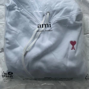 Vit hoodie från Ami Alexandre Mattiussi - Säljer en stilren vit hoodie från Ami Alexandre Mattiussi med röd hjärtlogga.Tröjan har luva med snören och är tillverkad i mjuk bomull. Jag har själv aldrig haft på mig den och den är storlek S men det finns exakt likadan i storlek M