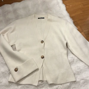 Vit kofta från Gina Tricot med knappar - Snygg vit kofta från Gina Tricot i storlek M. Koftan har v-ringning, stora bruna knappar framtill och på ärmsluten. Materialet är mjukt och känns stickat, perfekt för lager-på-lager. Passformen är normal och ärmarna är långa. I storleken är den rätt liten så en S kan även ha den 