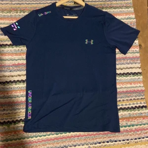 Blå Under Armour t-shirt, storlek  - Säljer en snygg marinblå t-shirt från Under Armour i storlek L. T-shirten har korta ärmar, coola tryckdetaljer i neonfärger och logga på bröstet. Materialet är lätt polyester som andas, perfekt för träning eller när du vill vara bekväm. Regular fit.