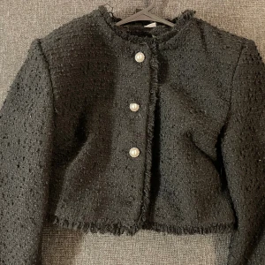 Jacka - Säljer en svart cardigan jacka i grovstickat material med fransad kant och tre snygga pärlknappar framtill. Jackan är croppad och har lång ärm, perfekt för att ge din outfit en chic touch.