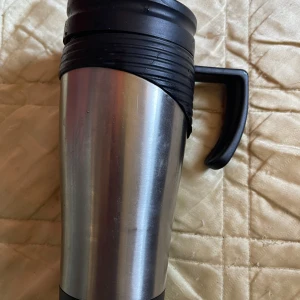 Termosmugg i rostfritt stål - Praktisk termosmugg i rostfritt stål med svart plastlock och handtag. Perfekt för kaffe eller te på språng. Muggen  är i gott bruksskick. Locket är avtagbart och har en öppning för att dricka direkt ur muggen.