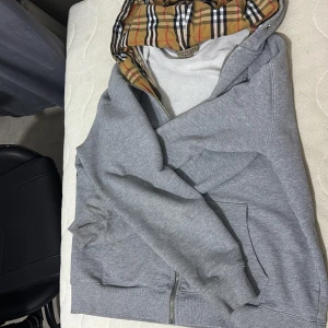 Grå Burberry hoodie  - Säljer en grå hoodie från Burberry med klassiskt rutigt foder i huvan. Tröjan har dragkedja, broderad logga på bröstet och känguruficka. Perfekt för dig som vill ha en stilren och lyxig streetwear-look.