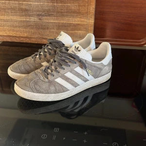 Adidas Gazelle grå/vita sneakers - Säljer ett par klassiska Adidas Gazelle sneakers i grå mocka med vita detaljer och de ikoniska tre ränderna på sidorna. Skorna har vit plös och häl, samt grå snörning och platt sula. Perfekta för dig som gillar retrostil och streetwear.