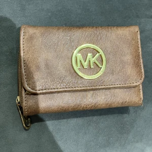 Brun plånbok  - Snygg brun plånbok med guldigt. Plånboken är tillverkad i skinn och har en praktisk dragkedja på sidan. Perfekt storlek för att ha med sig överallt och passar dig som gillar stilrena accessoarer.