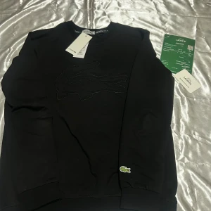 Svart sweatshirt från Lacoste med broderad logga - Svart sweatshirt från Lacoste med stor broderad krokodil på bröstet och broderad Lacoste-text längs ena ärmen. Klassisk rund hals och ribbade muddar. Liten grön Lacoste-logga vid nederkanten. Mjuk bomullskvalitet, perfekt för chill och streetwear.