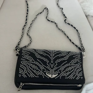 Svart axelväska med silverdetaljer - Cool svart axelväska med silverfärgade nitar i ett mönster på framsidan och en snygg kedja med inslag av svart läder. Väskan har dragkedja både fram och bak samt ett emblem med vingar i silver. Perfekt storlek för det viktigaste och ger en edgy vibe.
