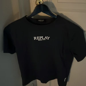 Svart t-shirt från Replay - Svart t-shirt från Replay med vit logga 'REPLAY SINCE 1981' tryckt på bröstet. Klassisk rund halsringning och korta ärmar. T-shirten har en enkel och stilren design som passar perfekt till jeans eller shorts.