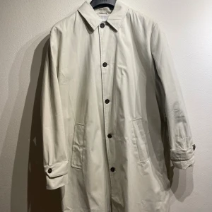 Beige trenchcoat rock - Elegant ljusbeige trenchcoat med klassisk krage och knäppning framtill. Den har praktiska fickor och en unik detalj med ett rutigt mönster på övre delen av ryggen. Perfekt för en stilfull och sofistikerad look.