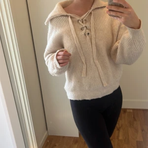 Beige stickad tröja med snörning - Supermysig beige stickad tröja från Anele med bred krage och snygg snörning framtill. Tröjan har en loose passform och långa ärmar, perfekt för lager på lager. Ribbstickad struktur som ger en chill och trendig vibe.