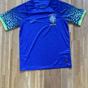 Blå Brasilien fotbollströja Nike - Snygg blå Brasilien fotbollströja från Nike med grönt Nike-märke och CBF-logga på bröstet. Ärmarna har leopardmönster i blått och grönt med gula muddar. Tillverkad i lätt och ventilerande material, perfekt för match eller träning.