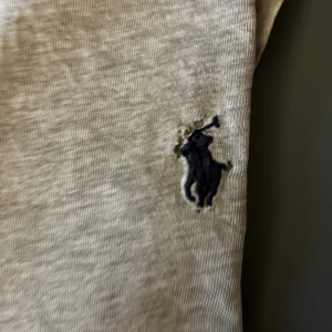 Grå t-shirt från Polo Ralph Lauren - Snygg grå t-shirt från Polo Ralph Lauren med klassisk marinblå broderad logga på bröstet. T-shirten har rund halsringning och är tillverkad i mjuk bomull för en skön känsla. Perfekt basic-plagg med stilren design. Litet hål vid loggan därför priset