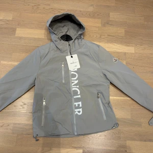 Grå vindjacka från Moncler med huva - Snygg grå vindjacka från Moncler med stor logga framtill, justerbar huva och tre dragkedjefickor. Jackan har långa ärmar, elastiska muddar och Moncler-märke på ärmen. Perfekt för dig som vill ha en stilren och sportig look.