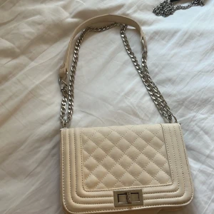 Beige quiltad axelväska med kedja - Stilren beige axelväska med quiltad framsida och silverfärgad kedjerem. Väskan har ett klassiskt rektangulärt format, vridlås i metall och invändigt fack med dragkedja. Perfekt för dig som gillar en chic och modern look.