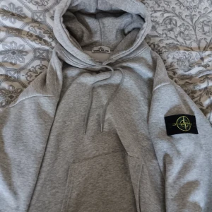 Grå hoodie från Stone Island - Säljer en grå hoodie från Stone Island med klassisk logga på ärmen. Tröjan har huva med snörning och en stor magficka framtill. Perfekt för dig som gillar streetwear och vill ha något stilrent men ändå med attityd.