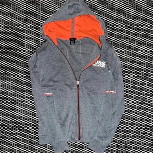 Grå hoodie från Hugo Boss  - Snygg grå hoodie från Hugo Boss med vit logga på bröstet och coola ränder i bokstäverna. Tröjan har orange foder i huvan, hel dragkedja och långa ärmar. Perfekt för dig som gillar stilren streetwear med en twist.