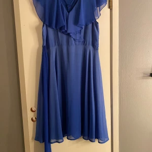 Blå långklänning med volang från Isle - Snygg blå långklänning från Isle med volangdetaljer över axlarna och v-ringning framtill. Klänningen har ett brett midjeband och är tillverkad i ett lätt, flödande syntetmaterial som ger en elegant siluett. Perfekt för dig som gillar färg och volym.💙