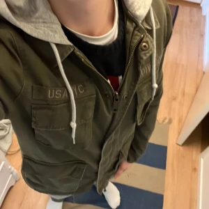 Khakigrön field jacka med grå mjukis hoodie - Säljer en field jacka i khakigrön färg med grå huva i sweatshirtmaterial. Jackan har fyra stora fickor framtill och dragkedja. Snygga detaljer och jackan är i väldigt fint skick, storlek S men passar även M. Hör av dig vid några frågor och priset är diskuterbart skriv till mig innan du köper så kan jag lösa fri frakt!
