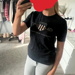 Svart t-shirt från Gant med tryck - Svart t-shirt från Gant med klassiskt tryck och sköld på bröstet. Modellen är normal i passformen och har rund halsringning samt korta ärmar. Perfekt för en sportig och stilren look.