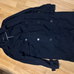 Svart trenchcoat från Vero Moda - Stilren svart trenchcoat från Vero Moda med klassisk krage, dubbelknäppning och bälte i midjan. Jackan har långa ärmar och är tillverkad i ett mjukt tyg som känns skönt mot kroppen. Perfekt för dig som vill ha en tidlös look.