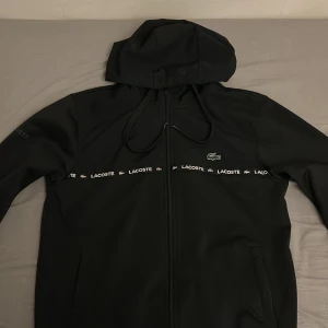 Svart vindjacka från Lacoste med huva - Svart vindjacka från Lacoste med huva och dragkedja. Jackan har logoband med Lacoste-tryck och krokodilemblem över bröstet. Regular fit och praktiska fickor framtill. Perfekt för dig som gillar stilren och sportig look.
