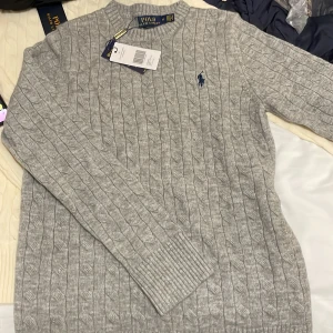 Grå kabelstickad tröja Polo Ralph Lauren - Snygg grå kabelstickad tröja från Polo Ralph Lauren med klassisk rund hals och marinblå broderad logga på bröstet. Tröjan har långa ärmar och ribbade muddar, perfekt för dig som gillar stilren och tidlös design.