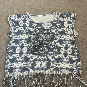 Blåvit tie-dye linnetopp med fransar - Cool linnetopp i blått och vitt tie-dye-mönster med stort svart tryck framtill. Toppen har fransad nederkant som ger en bohemisk vibe. Perfekt för sommardagar eller festivaler. Storlek S.