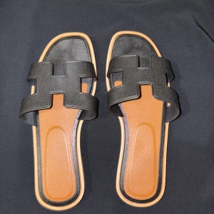 Svarta sandaler med H-design - Snygga svarta sandaler med breda remmar i skinn och cool H-formad design över foten. Sulan är platt och beige med svarta detaljer, och innersulan är brun. Perfekt för dig som gillar stilrena och trendiga skor med öppen tå och fyrkantig form.