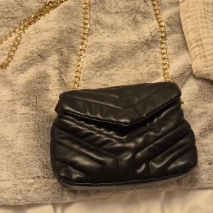 Svart quiltad axelväska med guldkedja - Snygg svart axelväska i quiltad design med guldfärgad kedja som axelrem. Väskan har ett lock med magnetlås och flera fack inuti för smart förvaring. Perfekt att ha mobil samt annat för ute kväll💕 Köpt för 150. Skriv om du har frågor!