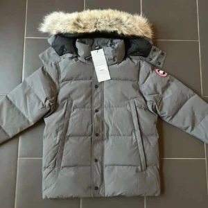 Grå dunjacka från - Säljer en grå dunjacka från  med pälskantad huva och klassisk logga på ärmen. Jackan har långa ärmar, dragkedja och knappar framtill samt två stora fickor. Perfekt för kalla vinterdagar och har en stilren, boxig passform.