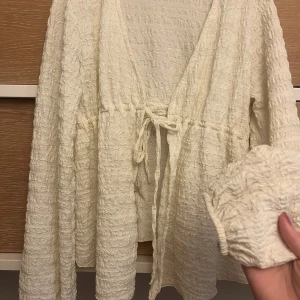 Krämvit omlottklänning från Gina Tricot - Säljer en krämvit omlottklänning från Gina Tricot i storlek XS. Klänningen har lång ärm, snygg struktur med rynkad effekt och knyt i midjan. Perfekt för dig som gillar en feminin och chill vibe. Materialet känns mjukt och har en lätt textur.