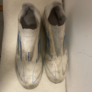 Adidas F50 vita och blå fotbollsskor - Säljer ett par Adidas F50 fotbollsskor i storlek 40. Skorna är vita med blå detaljer och har en strömlinjeformad design utan snörning. Yttermaterialet är syntet och sulan har dobbar för spel på gräs. Perfekt för dig som vill ha snabba och lätta skor på planen. Priset diskuteras vid snabb affär. 