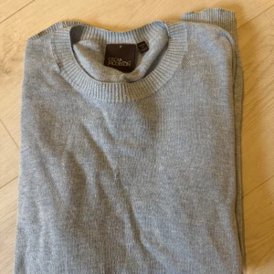 Ljusblå stickad tröja Oscar Jacobson - Snygg ljusgrå stickad tröja från Oscar Jacobson. Klassisk rund halsringning, ribbade muddar och nederkant. Perfekt för lager-på-lager och enkel att matcha med jeans eller chinos. Tillverkad i mjukt material för en skön känsla.