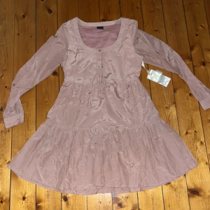 Rosa klänning från Gina Tricot - Supersöt ljusrosa klänning från Gina Tricot i modellen Cassandra Dress. Klänningen har lång ärm, rund halsringning och knappar framtill. Tillverkad i en lyxig mix av bomull och silke som ger ett fint skimmer. Volang nertill för extra flow.