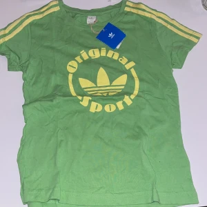 Grön Adidas t-shirt med gula detaljer - Snygg grön t-shirt från Adidas med gula ränder på axlarna och stort gult tryck med Adidas-logga och texten 'Original Sport' på bröstet. Klassisk passform och rund halsringning. Perfekt för en sportig look.