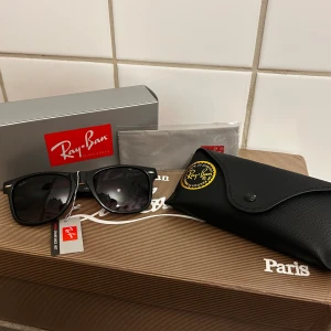vinröda Ray-Ban Wayfarer solglasögon - Klassiska vinröda Ray-Ban Wayfarer solglasögon med mörka glas och ikonisk fyrkantig form. Kommer med original svart fodral i skinnimitation, putsduk och Ray-Ban box. Perfekt accessoar för en cool och tidlös look.
