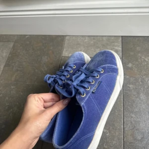 Blå sammetssneakers från Superga - Snygga blå sneakers från Superga i mjuk sammet med vita platta sulor och guldfärgade öljetter. Skorna har klassisk snörning och rund tå, vilket ger en cool och trendig vibe. Perfekta för dig som gillar att sticka ut med färg och material.