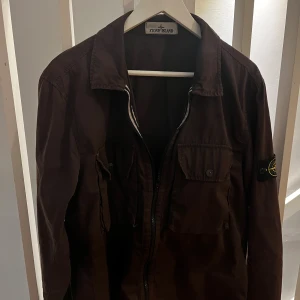Brun overshirt från Stone Island - Säljer en brun overshirt från Stone Island med klassisk krage, två bröstfickor med knappar och ikonisk patch på ärmen. Jackan har dragkedja framtill och är tillverkad i bomull. Perfekt för dig som gillar streetwear och snygga detaljer.