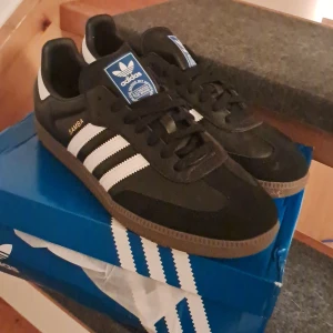 HELT NYA Adidas Samba svart/vit sneakers - Klassiska Adidas Samba sneakers i svart skinn och mocka med vita ränder och brun gummisula. Ikonisk design med blå Adidas-logga på plösen och guldfärgad 'SAMBA'-text på sidan. Perfekta för dig som gillar retrostil och streetwear.